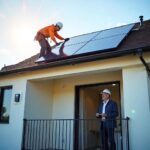 A rooftop scene during the final phase of a solar installation on a modern multi-family apartment building. Two professional PV technicians in safety gear secure the last panel in place, while below, the building owner watches from a balcony, smiling with visible relief and excitement. The sky is clear, the late afternoon sun reflects off the newly installed solar array. A subtle digital meter display on the wall near the entrance shows “System Online – Clean Energy Active.” The atmosphere captures the pride, professionalism, and optimism that comes with the completion of a sustainable energy system.